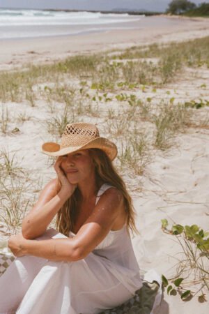 A woman sitting on the beach wearing a natural straw cowboy hat, with grass and ocean in the background.
