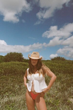 A woman wearing The Desert Cowboy straw fedora hat in natural color, standing outdoors with a green background and blue sky.