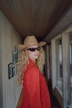 A woman wearing a tan straw cowboy hat with a desert rose weave detail, styled with sunglasses and a red jacket.