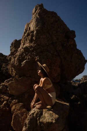 Model wearing the Mira Wide Boater hat, sitting on rocks by the ocean