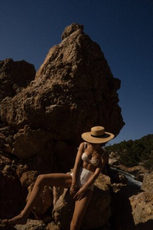 A woman wearing a Mira Wide Boater hat, sitting on rocks by the water, with a clear blue sky above.