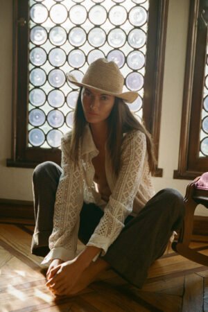 Model wearing a Raffia Cowboy straw hat, sitting on the floor in a sunlit room with patterned windows.