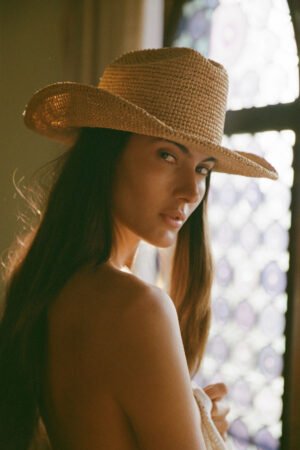 Model wearing a natural raffia cowboy hat, with a soft background and light filtering through a window.