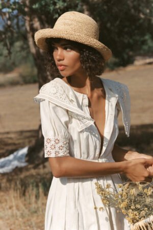 Model wearing a Raffia Cruiser straw boater hat in natural color, holding dried flowers.