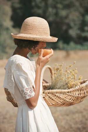 A woman wearing a natural straw boater hat, holding a basket with flowers and a drink.