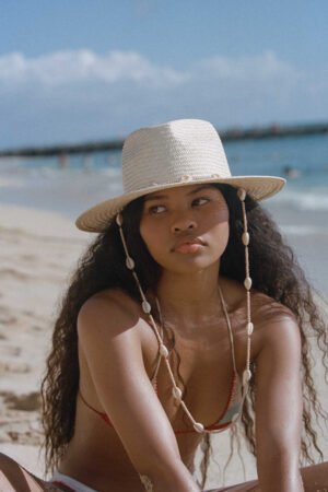 A woman wearing a straw Seashells Fedora hat with a chin strap, sitting on the beach.