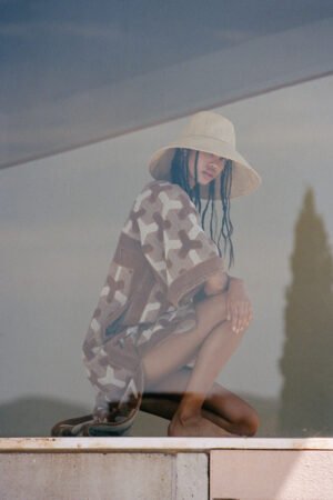 Model wearing The Cove straw bucket hat, crouching by a reflective surface, wearing a patterned cover-up.
