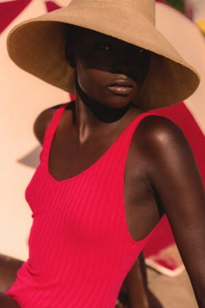 Model wearing The Cove straw bucket hat in natural color, seated on the beach, wearing a pink swimsuit.