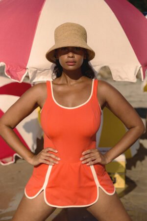 Model wearing The Inca Bucket straw hat, standing on the beach under an umbrella.