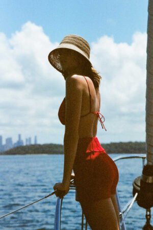 A woman wearing the Inca Bucket straw hat, standing on a boat by the water, with a city skyline in the background.