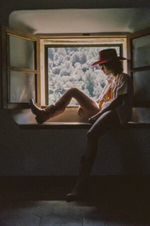 Model sitting by a window wearing a red wool felt cowboy hat and cowboy boots.