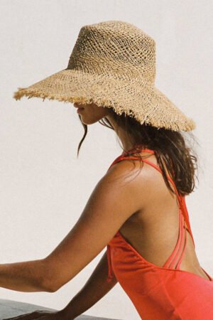 The Sundowner straw boater hat, natural color, worn by a model in a swimsuit, showcasing its wide brim and frayed edge.
