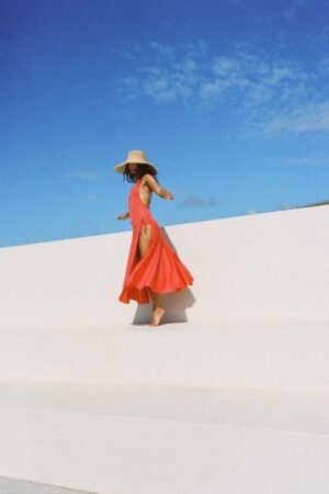 Model wearing The Sundowner straw boater hat in natural color, twirling on a white surface against a blue sky.