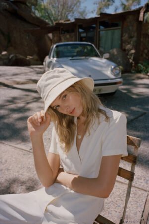 Model wearing a beige cotton bucket hat, seated outdoors with a car in the background.