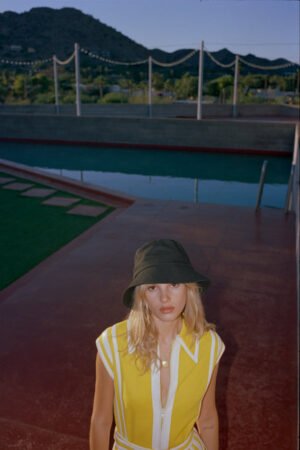 Model wearing a black cotton bucket hat, standing by a pool in a yellow outfit.