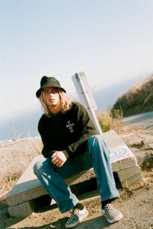 Young person wearing a black cotton bucket hat, sitting on a concrete block by a road with a scenic view.